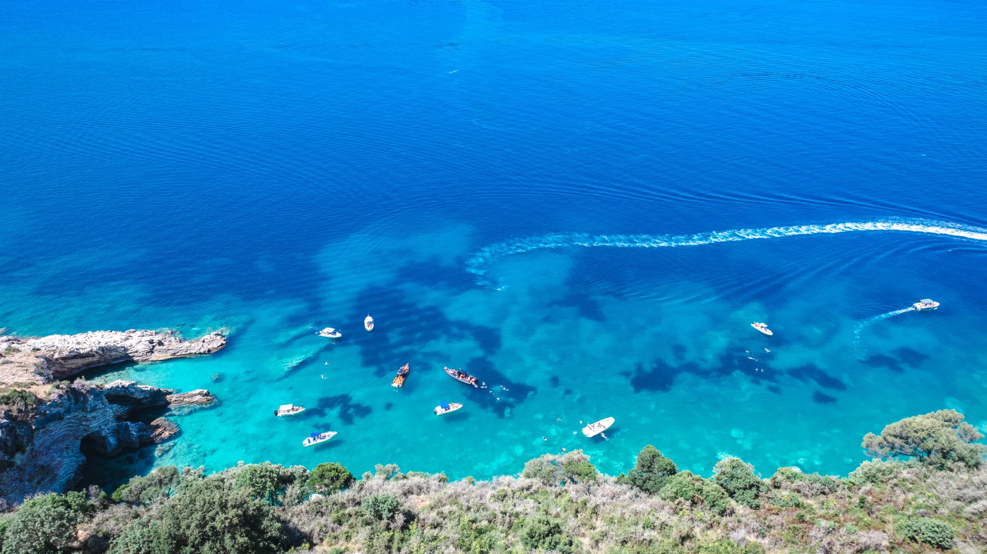 Aerial drone view of small boats anchored in a clear bay with one boat trailing a long wake
