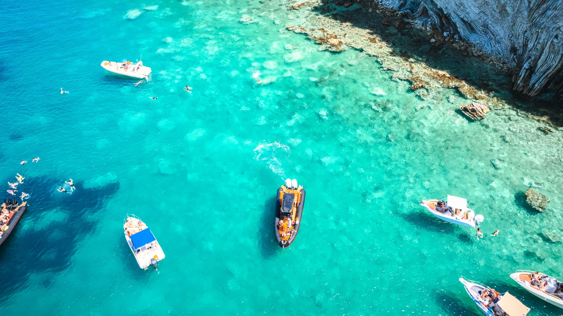 Aerial drone view of multiple boats anchored in shallow turquoise water near rocky Albanian Riviera coastline